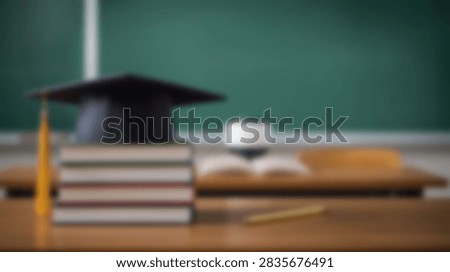 A graduation cap sits atop a stack of books on a wooden desk in front of a green blackboard. An open book and pencil are also visible.