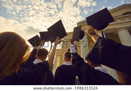 Graduation ceremony event celebration. Diverse student wearing gown mantles standing in group holding raised hat rear view from bottom. Shot over sunset and university high school building