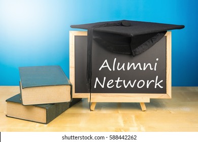 Graduation Hat, Books, Blackboard With Text Alumni Network On A Wooden Background.