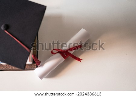 A graduation hat or mortarboard and diploma certificate paper tied with red ribbon on a stack of  books with empty space slightly undersaturated with vignette for vintage effect