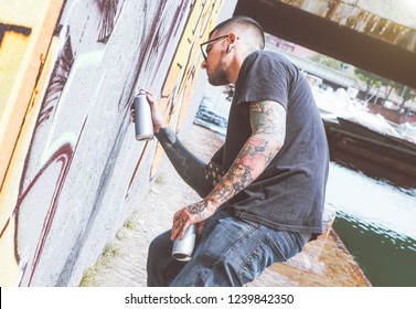 Graffiti Artist Painting With A Color Spray Can On The Wall In The City - Young Guy Doing A Mural Next To The River -  Urban And Street Art Lifestyle Concept