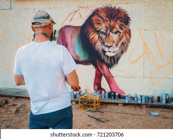 Graffiti Artist Painting On The Street Wall. Man With Aerosol Spray Bottle Near The Wall. Young Talented Guy In Protective Mask Drawing Colorful Lion.