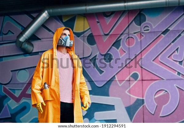Graffiti\
painter in respirator mask standing near the wall with his colorful\
paintings with copy space. Street art\
concept