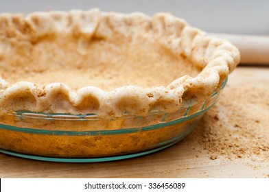 A Graham Cracker Coated Pie Crust Is In A Glass Pie Pan Waiting To Be Baked.  The Pie Dough Has A Fluted Edge.  Graham Cracker Crumbs Are Scattered Next To The Pie.