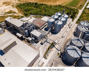A grain storage facility shows multiple silos and processing buildings in a rural area. Vehicles are parked nearby with fields visible in the background. The scene is captured from above.