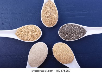 Grains Including, White Grain Quinoa, Psyllium Husk Powder, Black Chia, Wheat Germ, And Ground LSA Mix, In White Measuring Spoons On Dark Blue Wood Grain Table. 