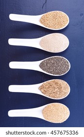 Grains Including, White Grain Quinoa, Psyllium Husk Powder, Black Chia, Wheat Germ, And Ground LSA Mix, In White Measuring Spoons On Dark Blue Wood Grain Table. 