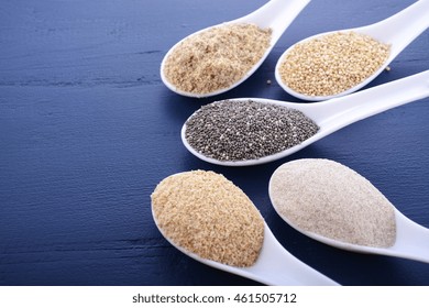 Grains Including, White Grain Quinoa, Psyllium Husk Powder, Black Chia, Wheat Germ, And Ground LSA Mix, In White Measuring Spoons On Dark Blue Wood Grain Table. 