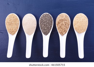 Grains Including, White Grain Quinoa, Psyllium Husk Powder, Black Chia, Wheat Germ, And Ground LSA Mix, In White Measuring Spoons On Dark Blue Wood Grain Table. 