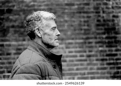 Grainy black and white analog film portrait of a contemplative senior man in profile against a blurred brick wall background, evoking a vintage, timeless feel - Powered by Shutterstock