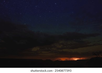 The Grand Canyon's silhouette is visible under a starry sky, with a distant glow suggesting a lightning storm or sunset on the horizon. - Powered by Shutterstock