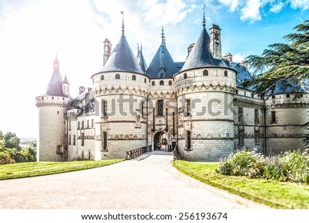 Grand castle in France, Europe. Chateau of white stone with towers surrounded with green lawns and trees. Road leading to entrance in foreground, blue cloudy sky in background. Architecture of Europe.