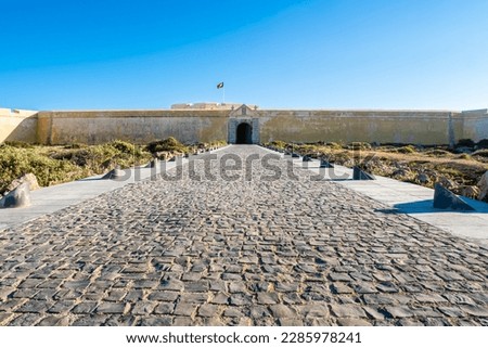 Grand entrance of Fortaleza de Sagres, a historic fortress in the Algarve area of Portugal, is captured in a stunning front view, with a vast driveway leading to its weathered walls and imposing gate.