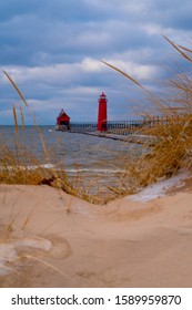 Grand Haven, MI—Dec 15, 2019; sand dunes with ice, snow and grass on top in foreground of red lighthouse and fog horn building marking entrance to Grand River shipping channel on Lake Michigan shore
