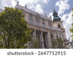 Grand historic building in Budapest featuring arched windows, ornate stonework, and a green domed tower with a golden spire, framed by trees and a flagpole.