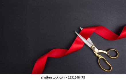 Grand opening. Top view of gold scissors cutting red silk ribbon against black background, copy space