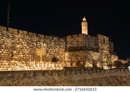 A grand stone wall, likely part of Jerusalem's Old City, is lit up at night, featuring a prominent, glowing menorah symbolizing the Hanukkah holiday