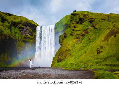 Grand Waterfall With Rainbow In The Water Mist Skogafoss. Elderly Woman Performs Asana 