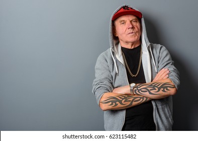 Grandad Transformed Into A Bold Teenager. Tattooed Arms Are Crossed, A Gold Chain Hangs Over A Black T-shirt. Big Empty Space For Inserting A Text Or A Picture On The Left Side Of The Photo.