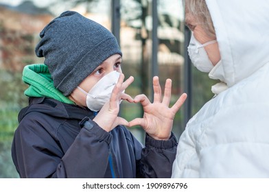 Grandchild with grandmother in respiratory mask outdoors in winter. Manifestation of feelings. Social distancing, contactless greeting. Coronavirus covid-19. Boy shows heart with his fingers. Love
