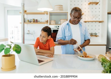 Grandchild and grandpa cooking together. The kid is looking at the laptop and checking on the recipe while grandad standing next to the stove and preparing a meal.