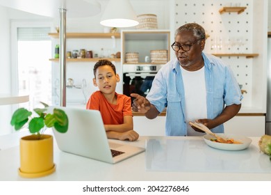 Grandchild And Grandpa Cooking Together. The Kid Is Looking At The Laptop And Checking On The Recipe While Grandad Standing Next To The Stove And Preparing A Meal.
