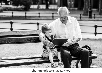 Grandchild Teaching To His Grandfather To Use Tablet On A Bench. Black And White Photography