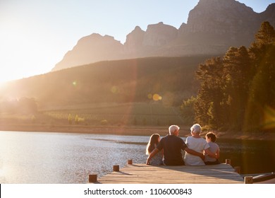 Grandchildren With Grandparents Sitting On Wooden Jetty By Lake