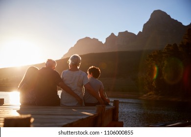 Grandchildren With Grandparents Sitting On Wooden Jetty By Lake
