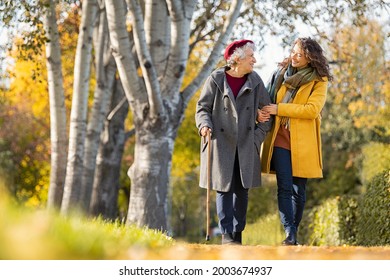 Granddaughter walking with senior woman in park wearing winter clothing. Old grandmother with walking cane walking with lovely caregiver girl. Happy woman and smiling grandma walking in autumn park.