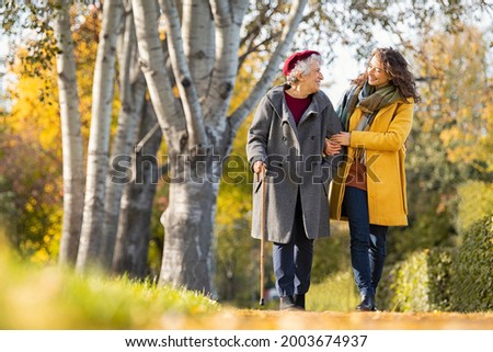 Granddaughter walking with senior woman in park wearing winter clothing. Old grandmother with walking cane walking with lovely caregiver girl. Happy woman and smiling grandma walking in autumn park.