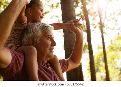 Grandfather giving granddaughter piggyback ride in park. Granddaughter sitting on shoulder of older man. 