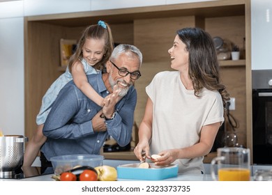 Grandfather giving his granddaughter a piggy back ride while the young woman makes lunch and laughs at them - Powered by Shutterstock