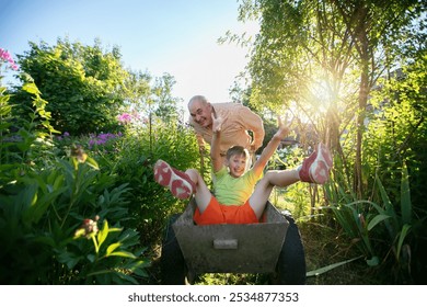 Grandfather giving his grandson a ride in a garden wheelbarrow on a summer day. Happy summer holidays. Family relaxing in the garden. - Powered by Shutterstock