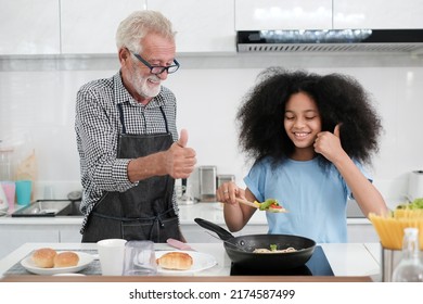 Grandfather And Granddaughter Making Food. Portrait Of Nice Cheerful Kind Grey-haired Grandparent Teaching Grandchild Cooking Homemade. At Modern Light White Interior Kitchen House. Spaghetti.