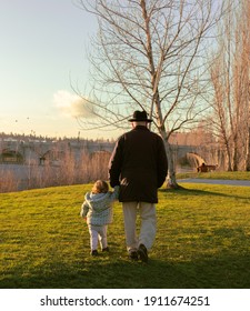 Grandfather And Granddaughter. Old Man And Little Girl Walking Backwards In The Park At Sunset With Grass And Bridge