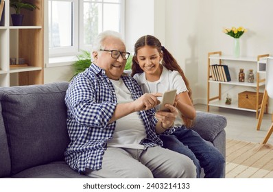 Grandfather and granddaughter spending time together while watching videos or photos on mobile phone. Old man and child sitting on sofa and looking at smartphone screen. Generations and technologies - Powered by Shutterstock