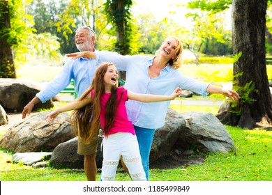 Grandfather And Grandmother Of The Elder Family With Granddaughter Relaxing Happiness And Dancing Joyfully On Green Grass In The Park.