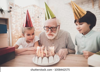 Grandfather, grandson and granddaughter at table at home. Grandpa is blowing candles on birthday cake.
