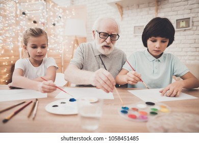 Grandfather, Grandson And Granddaughter At Table At Home. Children Are Painting.