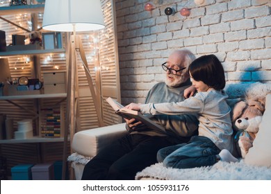 Grandfather and grandson on couch at night at home. Granddad is reading fairy tales book.