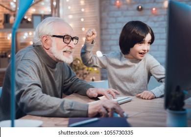 Grandfather And Grandson Are Playing Games On Computer At Table At Night At Home. Boy Is Cheering For Granddad.