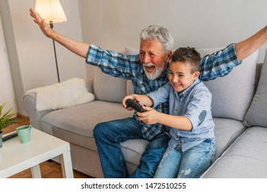 Grandfather and grandson playing video game with console, happy time together. Grandfather with his nephew seated on a sofa playing video games. Grandfather and child playing game console - Shutterstock ID 1472510255