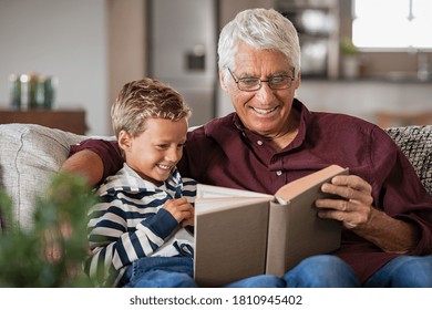 Grandfather And Grandson Reading A Book And Smiling. Happy Little Boy With Old Grandpa Reading Story Book At Home. Senior Man Tells A Fairy Tale Story To His Grandchild While Sitting On Couch.