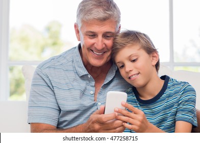 Grandfather And Grandson Using Mobile Phone At Home. Boy Helping Senior Man Surfing Net With Smart Phone. Grandfather And Grandchild Checking Their Photos On New Phone.
