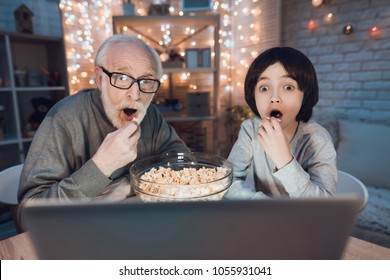 Grandfather and grandson are watching movie with popcorn at table at night at home.