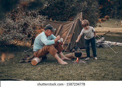 Grandfather and his grand child are camping, talking and eating canned  food. Boy is  stirring something is old сauldron. Image with selective focus and toning - Powered by Shutterstock