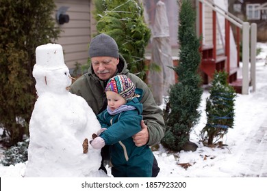 Grandfather And His Small Granddaughter Making Snowman On The Backyard Of Their Country House. Family Multi-generation Winter Leisure Activities.