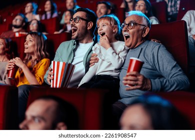 A grandfather and his son accompanied with his granddaughter are in the movie theater watching and exciting film together while enjoying beverages and popcorn.