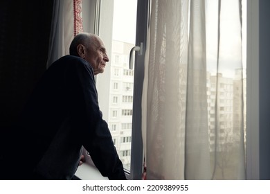 Grandfather Looking Through Window Glass Covered With Transparent White Curtain. Senior Man Waiting Guests With Hope.